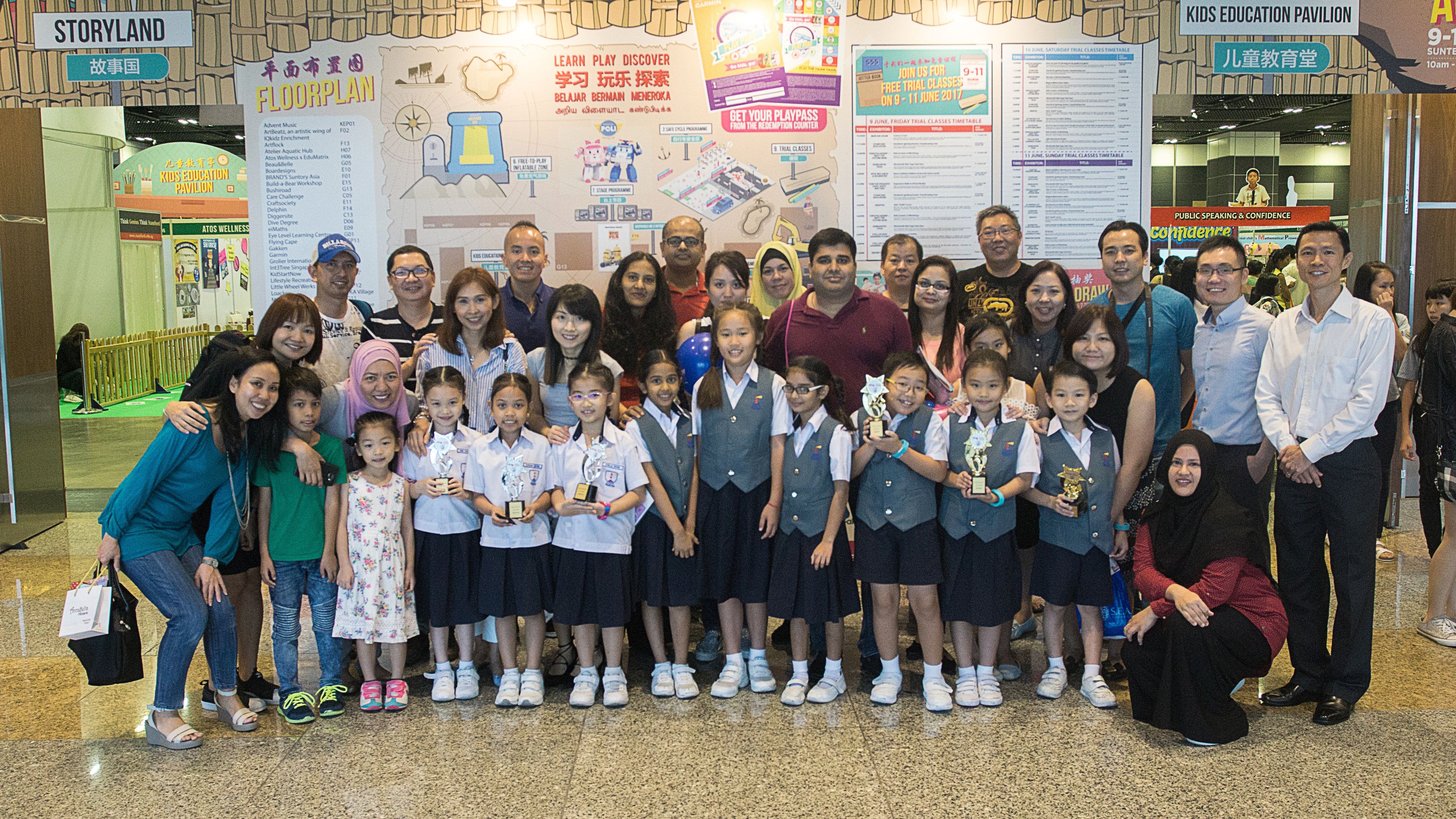 A large group of adults and uniformed schoolgirls holding trophies pose together at a Kids Education Pavilion event.